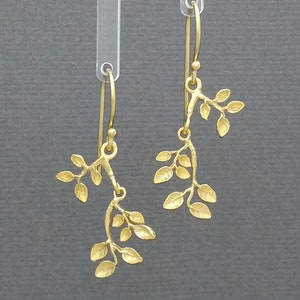 May include: A pair of gold dangle earrings featuring a delicate branch design with multiple leaves. The earrings are made of a smooth, textured metal and have a simple, elegant style.