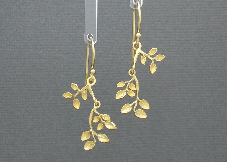 May include: A pair of gold dangle earrings featuring a delicate branch design with multiple leaves. The earrings are made of a smooth, textured metal and have a simple, elegant style.