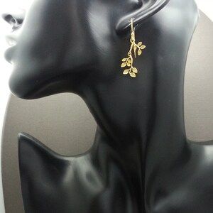 May include: Gold dangle earrings with a delicate leaf design. The earrings are shown on a black mannequin head.