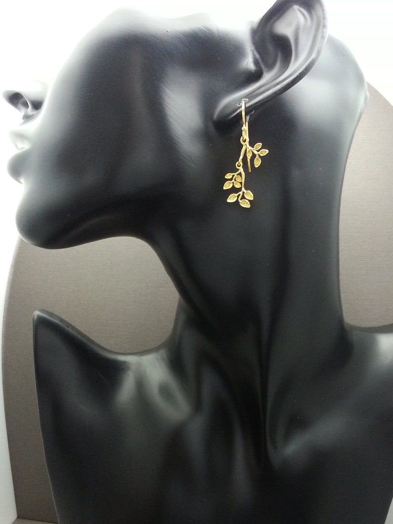 May include: Gold dangle earrings with a delicate leaf design. The earrings are shown on a black mannequin head.