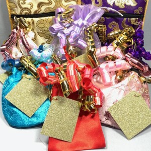 May include: Five small, colourful gift bags with gold, purple, pink, blue, and red fabric. Each bag has a gold tag attached and is decorated with ribbons and bows in various colours.
