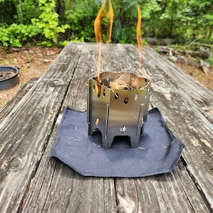 May include: A small, stainless steel camping stove with flames rising from the top. The stove sits on a dark blue, rectangular mat on a weathered wooden picnic table. A small fire pit is in the background.