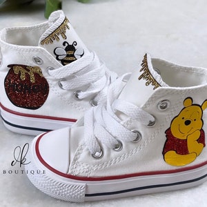 May include: White high-top sneakers with a Winnie the Pooh design. The shoes feature a honey pot, a bee, and Winnie the Pooh with glitter accents.