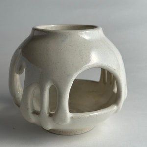 May include: White ceramic candle holder with a unique dripping design and open sides. The holder has a round shape and a smooth, glossy finish.