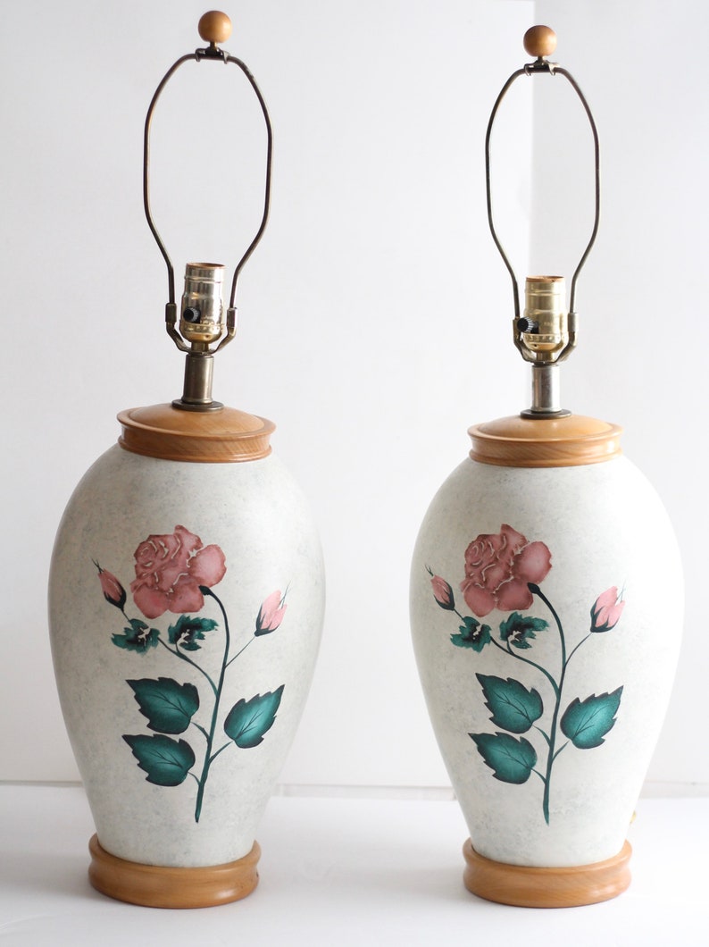 May include: A pair of ceramic table lamps with a white base and a wooden top. Each lamp features a hand-painted pink rose with green leaves.