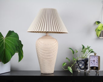 May include: A cream-coloured ceramic table lamp with a pleated white lampshade. The lamp base has a textured design with a series of raised circles.