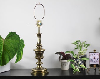 May include: A gold table lamp with a classic design and a white lampshade. The lamp is on a black surface with a green plant in a white pot and a clock on a stand.