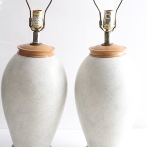 May include: A pair of white ceramic table lamps with wooden bases and gold metal lampshades. The lamps have a speckled finish and are wired with cords and plugs.