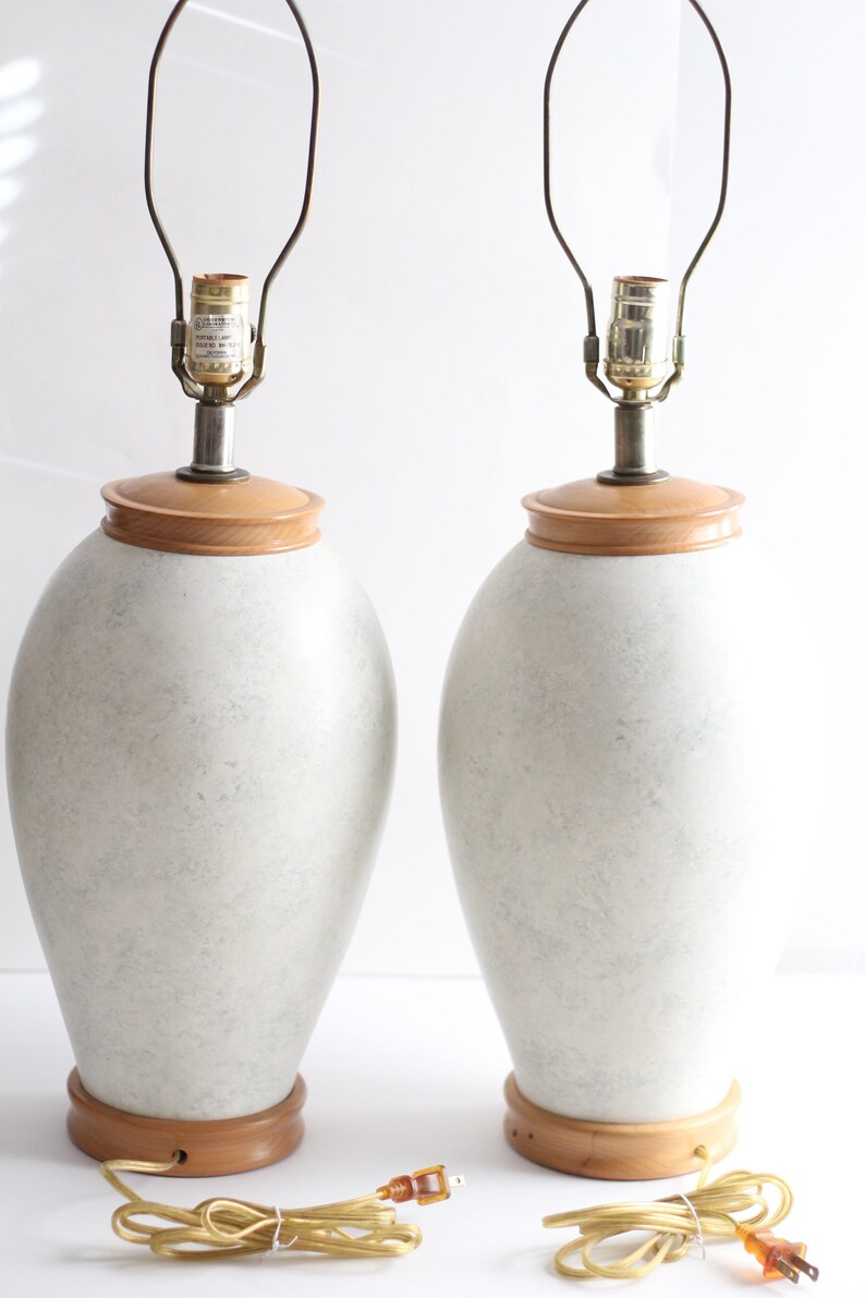 May include: A pair of white ceramic table lamps with wooden bases and gold metal lampshades. The lamps have a speckled finish and are wired with cords and plugs.