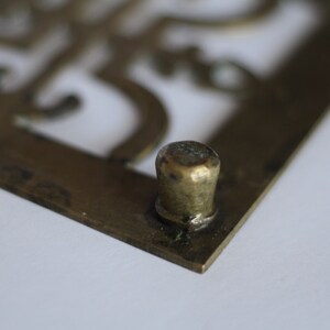 Op de afbeelding: Een vierkant decoratief object van messing met een uitgesneden ontwerp en een ronde messing knop op een hoek.