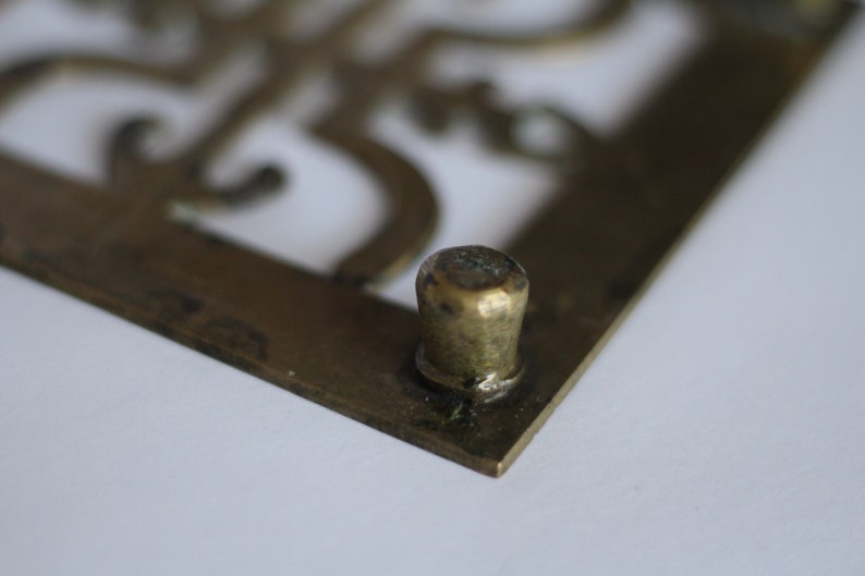 Op de afbeelding: Een vierkant decoratief object van messing met een uitgesneden ontwerp en een ronde messing knop op een hoek.
