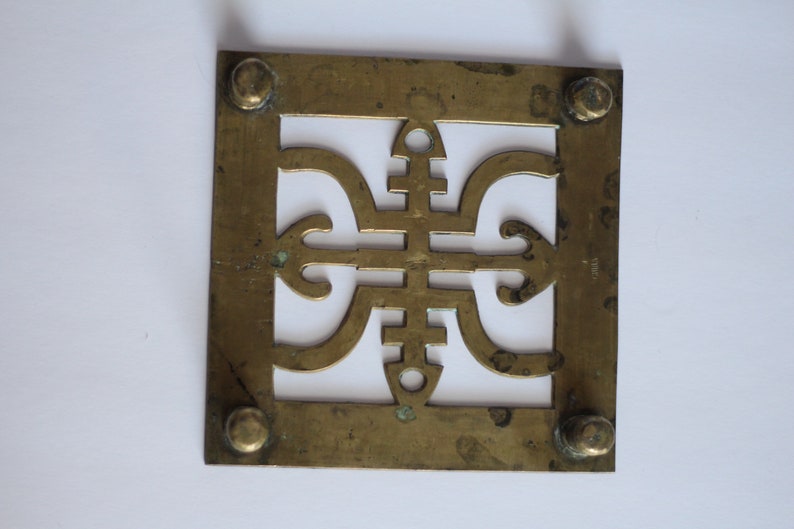 Op de afbeelding: Een vierkante messing plaat met een decoratief uitgesneden ontwerp. De plaat heeft vier kleine ronde knoppen op de hoeken.