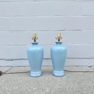 Peut inclure: Deux lampes de table en céramique bleu clair avec des accents dorés et une bordure blanche. Les lampes sont sur une surface en béton gris.