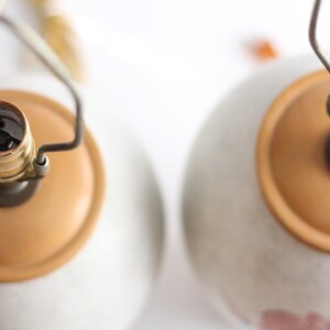 May include: Two ceramic table lamps with beige bases and gold-coloured light bulb sockets. The lamps have a simple design and are ready to be used.