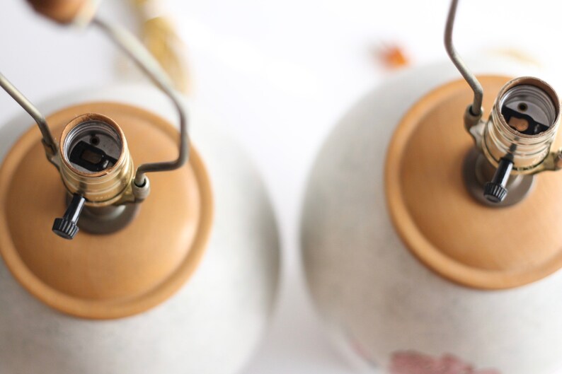 May include: Two ceramic table lamps with beige bases and gold-coloured light bulb sockets. The lamps have a simple design and are ready to be used.