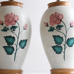 May include: A pair of white ceramic table lamps with a wooden base. Each lamp features a hand-painted design of a pink rose with green leaves.