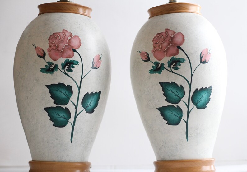 May include: A pair of white ceramic table lamps with a wooden base. Each lamp features a hand-painted design of a pink rose with green leaves.