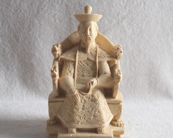 Op de afbeelding: Een wit keramiek beeldje van een man in traditionele Chinese kleding die op een troon zit. De troon is versierd met ingewikkelde houtsnijwerk van draken en andere mythische wezens.
