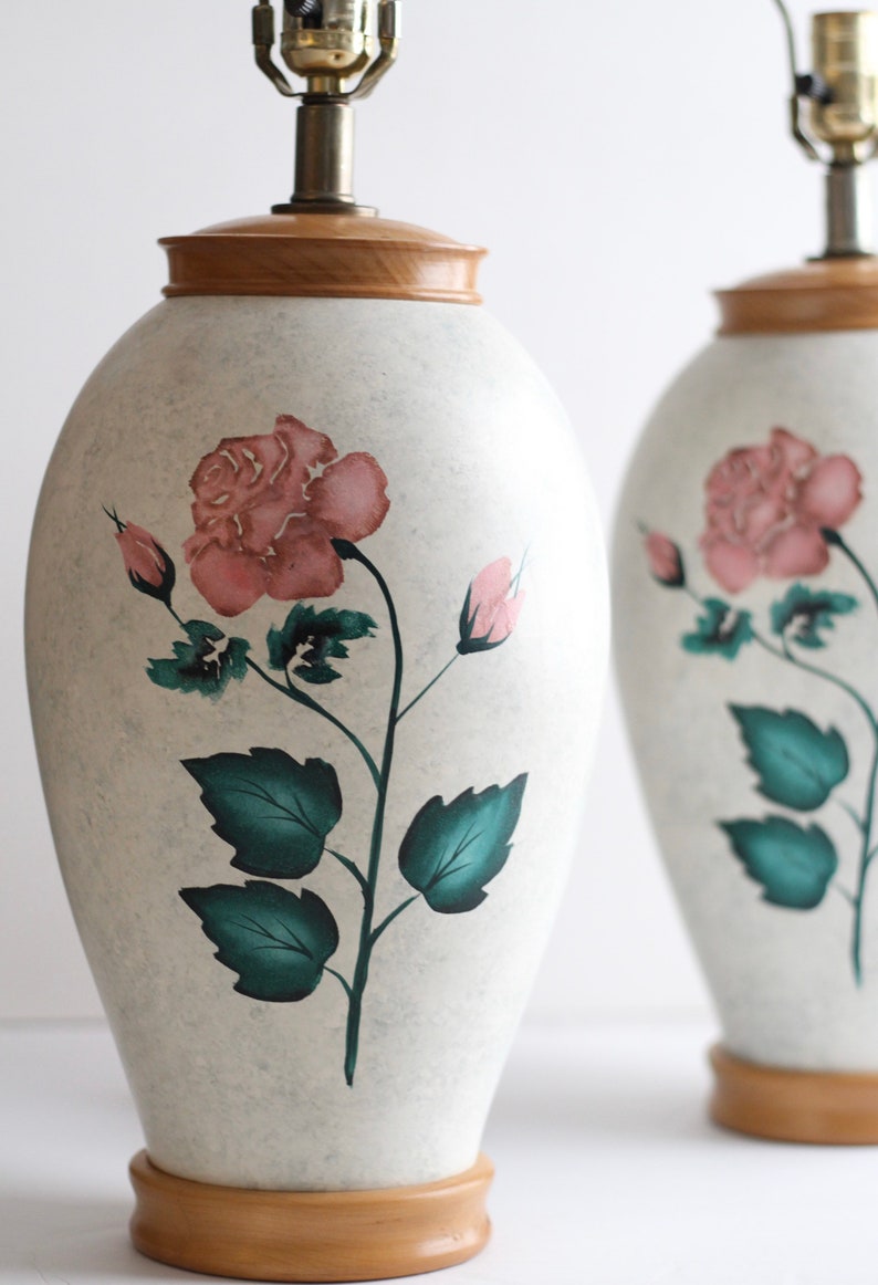 May include: Pair of ceramic table lamps with a white base and a wooden top. The lamps feature a hand-painted design of pink roses and green leaves.