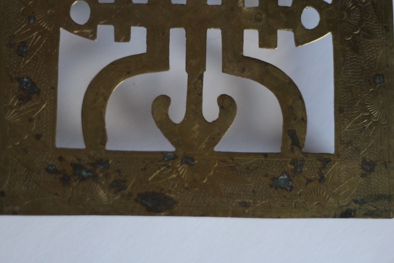 Op de afbeelding: Een vierkant decoratief item van messing met een ingewikkeld ontwerp met een gestileerd symbool en bloemmotieven. Het vierkant heeft een versleten, verweerde uitstraling.