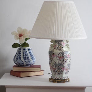May include: A white table lamp with a white pleated lampshade sits on a white table. The lamp base is a white ceramic vase with pink and gray floral designs. A blue and white ceramic vase with a flower sits on a stack of books on the table.