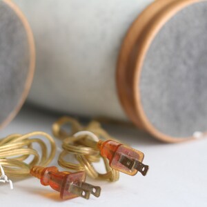 May include: Two gold electrical cords with orange plugs, coiled on a white surface. The cords are connected to two round, grey and white objects in the background.