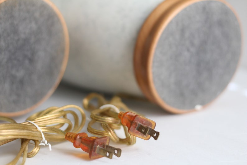 May include: Two gold electrical cords with orange plugs, coiled on a white surface. The cords are connected to two round, grey and white objects in the background.