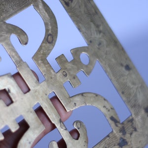 Op de afbeelding: Een decoratieve wandhanger van messing met een ingewikkeld uitgesneden ontwerp. Het ontwerp heeft een herhalend patroon van gestileerde bloemen en geometrische vormen. Het stuk heeft een verweerde, antieke afwerking.
