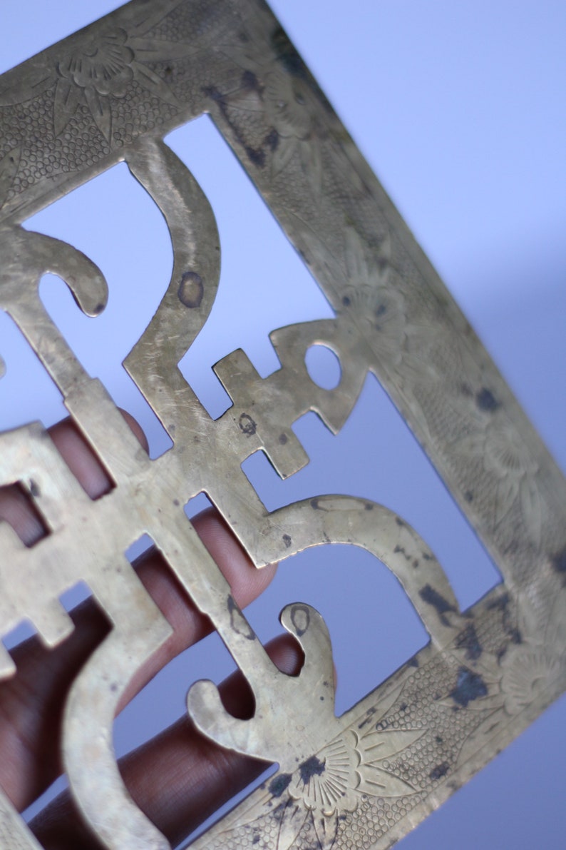 Op de afbeelding: Een decoratieve wandhanger van messing met een ingewikkeld uitgesneden ontwerp. Het ontwerp heeft een herhalend patroon van gestileerde bloemen en geometrische vormen. Het stuk heeft een verweerde, antieke afwerking.