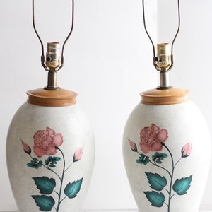 May include: Pair of ceramic table lamps with a white base and a pink rose design. The lamps have a wooden base and a brown wire lampshade.