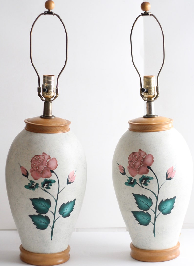 May include: Pair of ceramic table lamps with a white base and a pink rose design. The lamps have a wooden base and a brown wire lampshade.