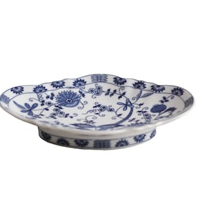 May include: A white ceramic serving dish with a blue floral pattern. The dish has a scalloped edge and a raised base.