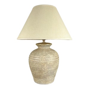 May include: A beige ceramic table lamp with a cream-coloured fabric lampshade. The lamp base has a textured, striated design.