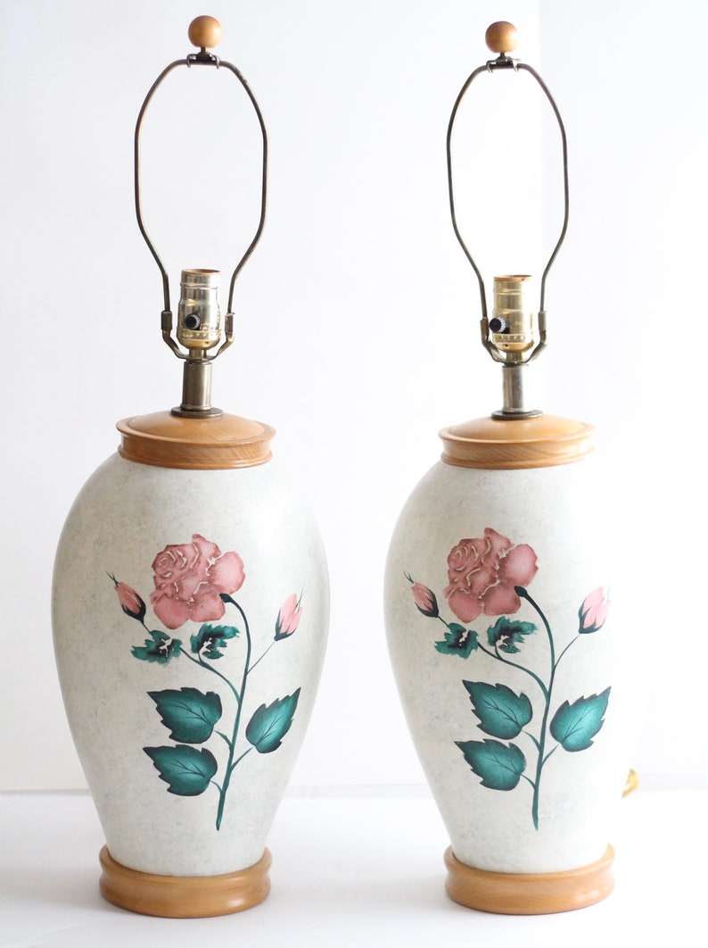 May include: A pair of ceramic table lamps with a white base and a pink rose design. The lamps have a wooden base and a brass lampshade.