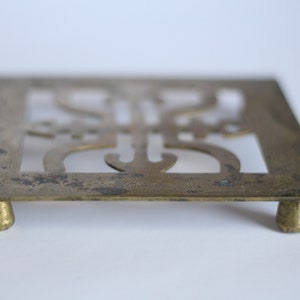 Op de afbeelding: Een messing trivet met een ingewikkeld ontwerp en vier poten. De trivet is vierkant en heeft een uitgesneden ontwerp in het midden.