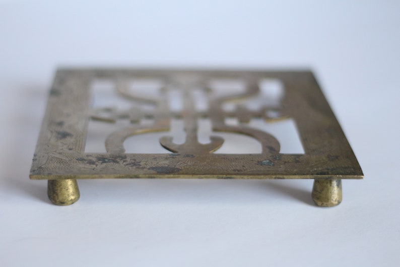 Op de afbeelding: Een messing trivet met een ingewikkeld ontwerp en vier poten. De trivet is vierkant en heeft een uitgesneden ontwerp in het midden.