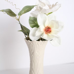 May include: A white ceramic vase with a textured, tree-like design holds a bouquet of white magnolia flowers with green leaves.