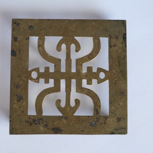 Op de afbeelding: Een vierkante messing trivet met een decoratief uitgesneden ontwerp. De trivet heeft een geometrisch patroon met een centraal motief dat lijkt op een gestileerde bloem of ster.