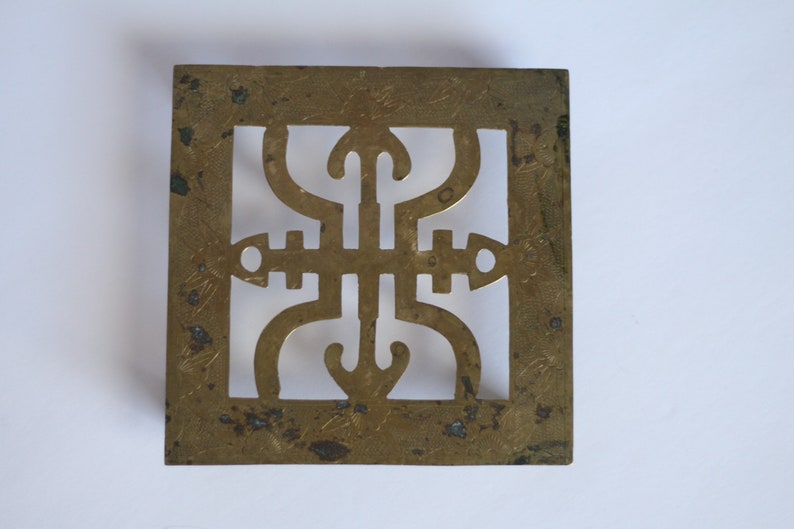 Op de afbeelding: Een vierkante messing trivet met een decoratief uitgesneden ontwerp. De trivet heeft een geometrisch patroon met een centraal motief dat lijkt op een gestileerde bloem of ster.