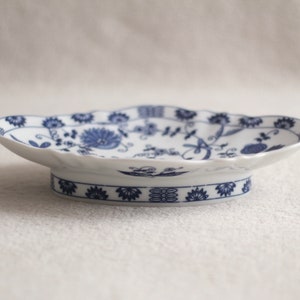 May include: A white porcelain serving dish with a blue floral pattern. The dish has a scalloped edge and a raised base.