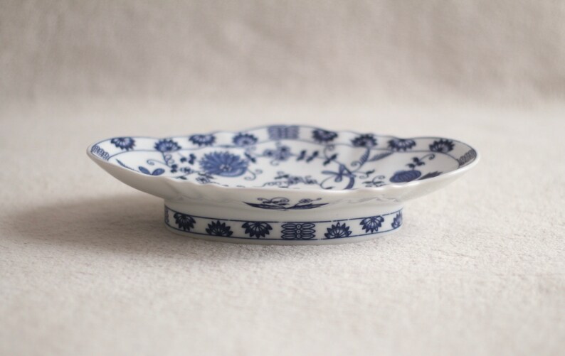 May include: A white porcelain serving dish with a blue floral pattern. The dish has a scalloped edge and a raised base.