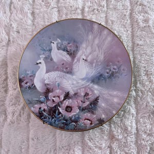 May include: A decorative plate featuring a painting of white peacocks and pink flowers. The plate has a gold rim and a soft, pastel color palette. The artwork includes three peacocks and several pink poppies.