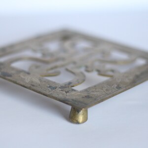 Op de afbeelding: Een zilverkleurige metalen onderzetter met een ingewikkeld, bloemig ontwerp. De onderzetter heeft vier poten en is vierkant.