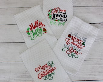 May include: Four white, textured Christmas kitchen towels with embroidered text and festive designs. The towels feature phrases like "Holly Jolly," "I've been good (ish) this year," "It's the Most Wonderful Time of the Year," and "Merry Christmas."