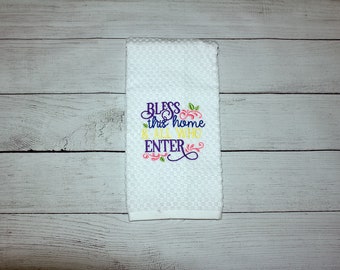 May include: White waffle-weave kitchen towel with embroidered text that reads "Bless this home & all who enter" in purple, yellow, and pink. The towel has a floral design and is displayed on a light wood surface.