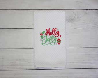 May include: White, textured hand towel with the embroidered words "Holly Jolly" in red and green. The design includes holly leaves, berries, and a Christmas ornament. The towel is displayed on a light wood surface.