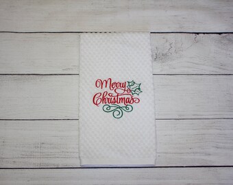 May include: White Christmas-themed hand towel with a textured, waffle-weave pattern. The words "Merry Christmas" are embroidered in red with green holly accents and flourishes. The towel is rectangular and displayed on a white wooden surface.