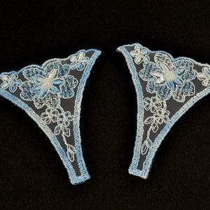 May include: Two light blue and white lace appliques with a floral design. The appliques are shaped like a triangle with a rounded top.