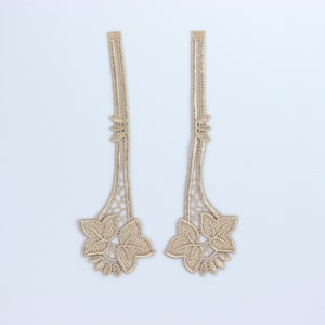 Puede incluir: Dos apliques de encaje de color dorado con un diseño floral. Cada aplique presenta una flor grande con cinco pétalos y hojas más pequeñas.