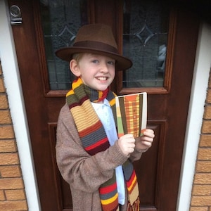 K&ouml;nnte beinhalten: Ein Kind tr&auml;gt einen braunen Fedora-Hut, einen langen, bunten Schal und eine braune Strickjacke. Der Schal hat Streifen in Rot, Gelb, Blau und Beige. Das Kind h&auml;lt ein Buch mit einem Karomuster.